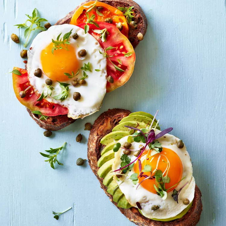 Rustic sourdough toast topped with fried eggs, heirloom tomatoes, avocado, capers, and micro-herbs on a soft blue background