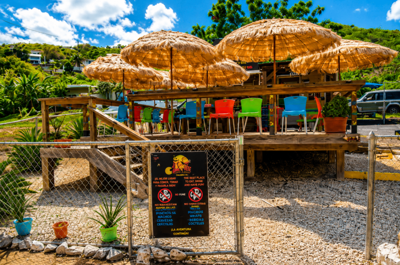 Come y Vete — open-air roadside kiosk with tiki umbrellas and bright plastic chairs