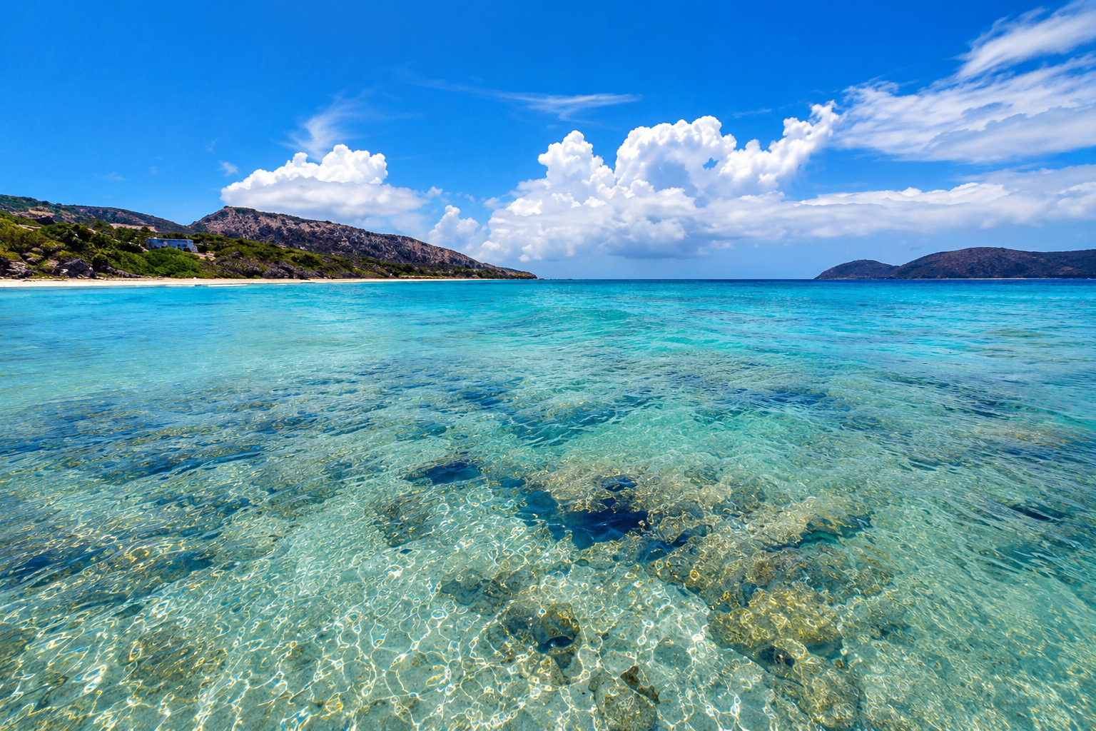 Crystal-clear turquoise shallows at Zoni Beach with coral visible beneath the water and hills in the distance